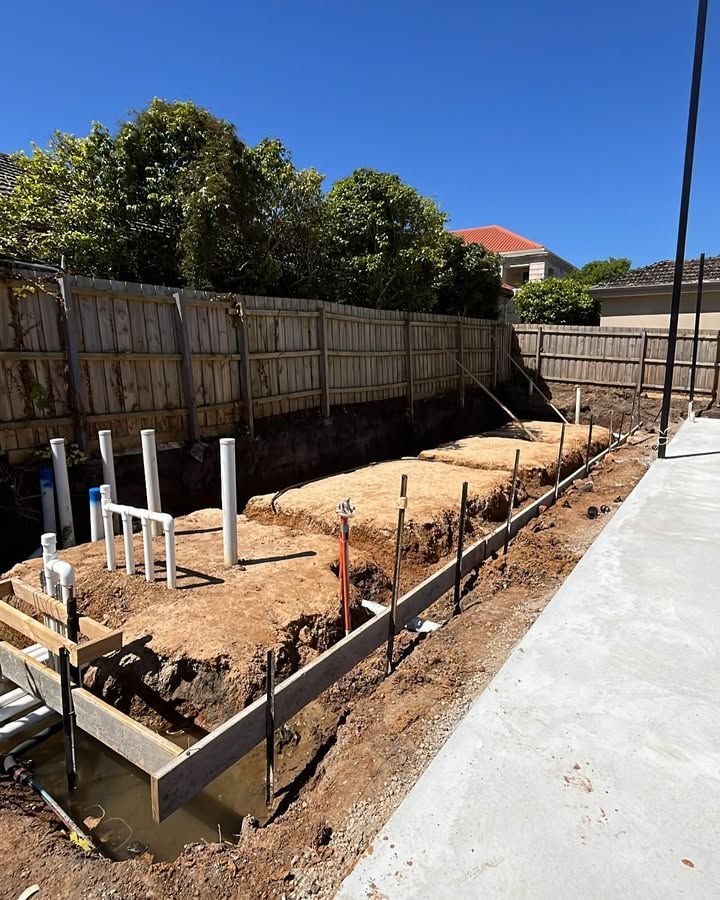 Timber formwork and plumbing pipes in footing trenches beside a fence