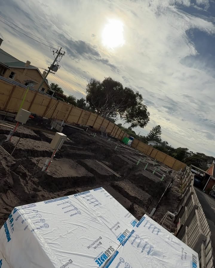 Footing trenches on a house site with wrapped slab pods in front, under a bright sky